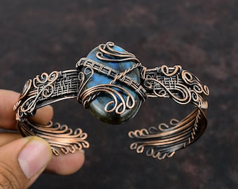 May include: A copper wire wrapped bracelet with a large oval labradorite gemstone. The bracelet has a woven design and intricate details.