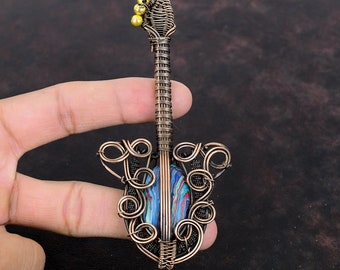 Puede incluir: Un colgante de guitarra de alambre de cobre con una incrustación de piedra de ágata de colores. La guitarra tiene un diseño de alambre detallado y una pequeña cuenta dorada en la parte superior del cabezal.