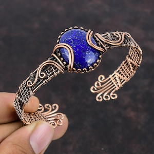 Puede incluir: Un brazalete envuelto en alambre de cobre con una piedra preciosa de lapislázuli azul. El brazalete tiene un diseño tejido y detalles intrincados.