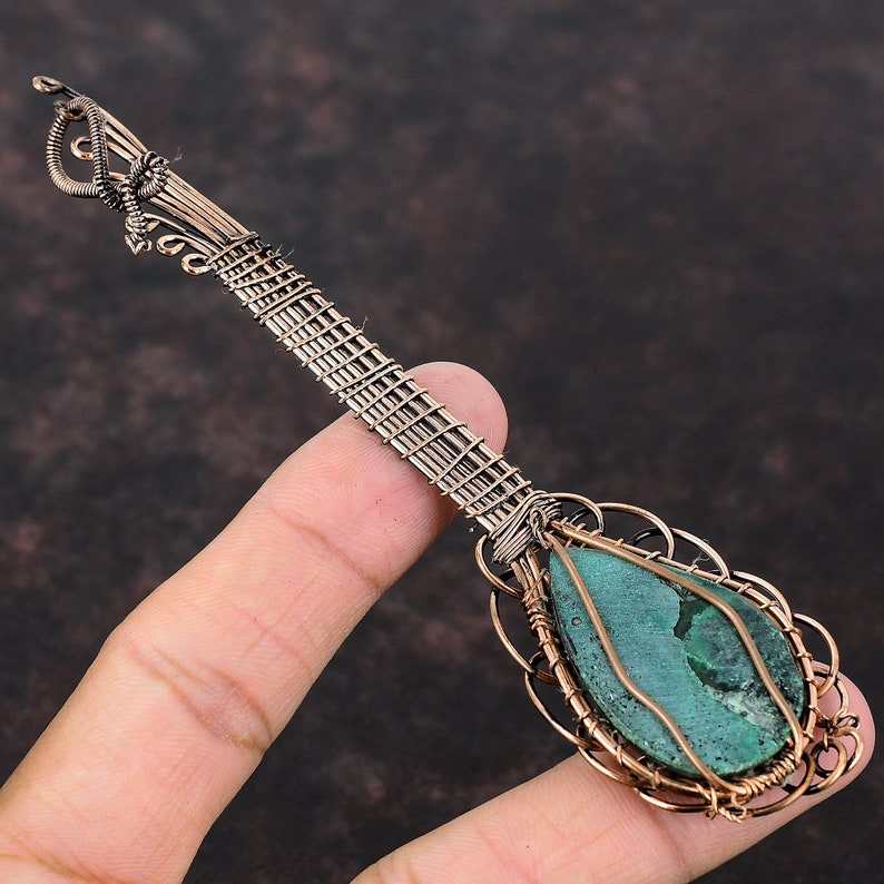 May include: A handcrafted copper-coloured pendant with a teardrop-shaped, teal-coloured stone. The stone is encased in a decorative wire frame, and the pendant has a long, intricately woven handle. The design is detailed and artistic.