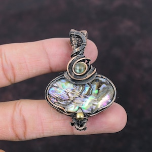 Op de afbeelding: Een hanger met koperdraad gewikkeld met een grote, iriserende abalone-schelp met een kleinere groene edelsteen aan de bovenkant.
