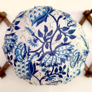 Op de afbeelding: Een rond, wit stoffen item met een blauw bloemenpatroon. Het ontwerp bevat grote, gestileerde bloemen en bladeren in verschillende tinten blauw. Het item lijkt handgemaakt.
