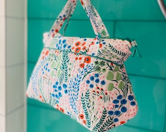 Op de afbeelding: Een witte, gewatteerde toilettas met een bloemenprint met blauwe, groene, oranje en roze bloemen. De tas heeft een turquoise rits en een turquoise stoffen handvat. De tas heeft een klein label met de tekst "caro" erop.
