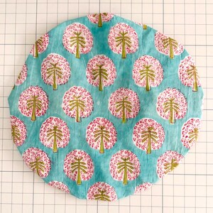 May include: A round, patterned fabric in shades of teal, pink, and green. The design features repeating tree-like motifs with pink, circular canopies and green trunks, set against a teal background. The fabric is likely cotton or a similar material.
