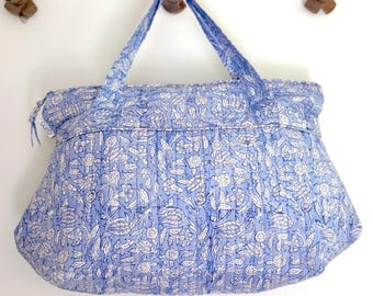 Op de afbeelding: Een lichtblauwe quilted tote bag met een wit bloemenpatroon. De tas heeft twee bovenste handvatten en een ritssluiting.