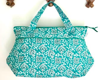 Op de afbeelding: Een tealblauwe tote bag met een wit bloemenpatroon. De tas heeft twee bovenste handvatten en een ritssluiting.