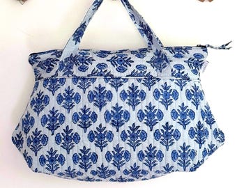 Op de afbeelding: Een blauwe en witte gewatteerde tote bag met een bloemenpatroon. De tas heeft een ritssluiting en twee bovenste handvatten.