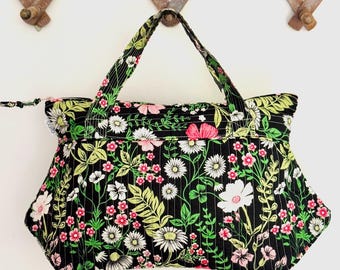 Op de afbeelding: Een zwarte draagtas met een bloemenpatroon van witte, roze en groene bloemen en bladeren. De tas heeft een ritssluiting en twee handvatten. De tas hangt aan een houten kledingrek.