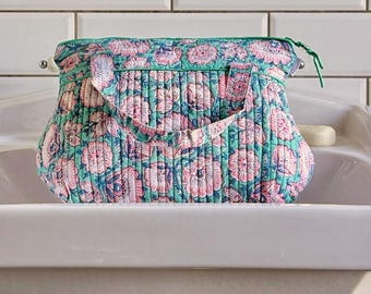 Op de afbeelding: Een gewatteerde tas met bloemenprint in tinten van teal, roze en blauw. De tas heeft een ritssluiting aan de bovenkant, een handvat en een bloemenpatroon. De tas staat op een witte wastafel.