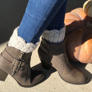 May include: A pair of brown ankle boots with a zipper and buckle detail. The boots are worn with a pair of light brown crocheted boot cuffs.
