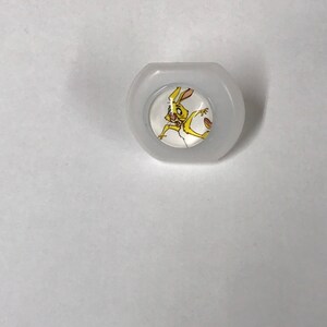 Op de afbeelding: Een witte plastic knop met een rond, doorzichtig midden met een cartoon afbeelding van een geel konijn met bruine ogen en een roze neus.