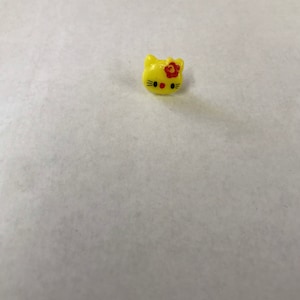 以下が含まれることがあります： 漫画の猫の顔と赤い花がついた小さな黄色のプラスチックボタン