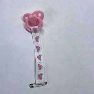 以下が含まれることがあります： 白いリボンが付いたピンクの哺乳瓶。リボンにはピンクの足跡模様と安全ピンが付いています。