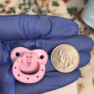 以下が含まれることがあります： ハート型のデザインで、漫画の豚が描かれたピンクの哺乳瓶。哺乳瓶は、サイズ比較のためにアメリカの25セント硬貨の隣にあります。