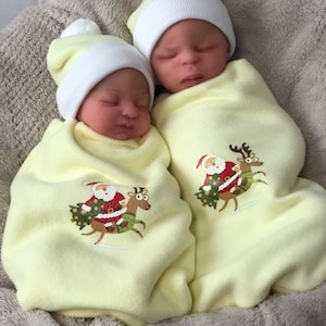 May include: Two baby dolls wearing yellow and white knit hats with pom-poms are swaddled in yellow blankets with Santa Claus and reindeer designs.