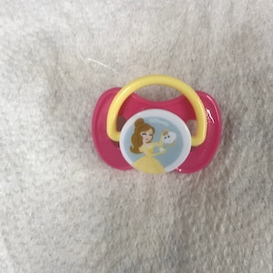 以下が含まれることがあります： ディズニーのプリンセスデザインが施されたピンクと黄色のベビー用おしゃぶり。美女と野獣のベルが描かれており、おしゃぶりには黄色の輪と、お茶碗を持ったベルが描かれた青と白のデザインがあります。