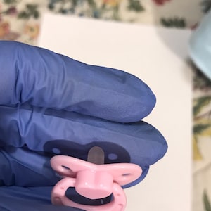以下が含まれることがあります： 透明な乳首が付いたピンク色のベビーおしゃぶり。おしゃぶりは、ハンドルとリング型のデザインが付いた丸みを帯びたシールドを備えています。おしゃぶりは青い手袋を着用した人が持っています。