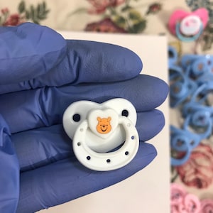 以下が含まれることがあります： ハート型のシールドに、くまのプーさんの漫画のデザインが描かれた白いおしゃぶり。おしゃぶりには白いリングと、縁の周りに複数の穴があります。