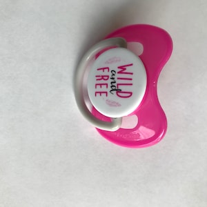 以下が含まれることがあります： ピンクのシリコン製おしゃぶりで、白い丸いディスクには黒い文字で「WILD and FREE」と書かれています。ディスクにはピンクの花柄のデザインが施されています。