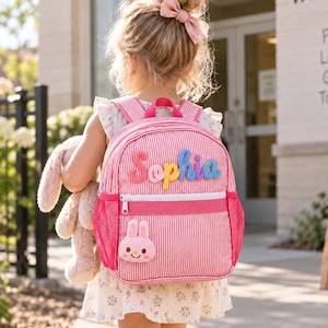 Puede incluir: Una mochila de rayas rosas y blancas con el nombre Sophia en letras coloridas y en relieve en el bolsillo delantero. La bolsa incluye una pequeña cara de conejo de peluche rosa en la parte inferior, una cremallera blanca y bolsillos laterales de malla rosa.