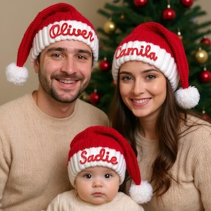 Könnte beinhalten: Eine Familie trägt passende rot-weiße Weihnachtsmützen mit personalisierten, rot gestickten Namen. Die Mützen haben weiße Bommel. Die Familie steht vor einem Weihnachtsbaum.