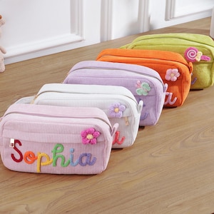 Puede incluir: Una fila de coloridos neceseres de pana. Las bolsas son en tonos rosa, blanco, morado, naranja y verde. La bolsa rosa tiene el nombre "Sophia" bordado en colores arcoíris. Cada bolsa tiene una flor o un adorno decorativo.
