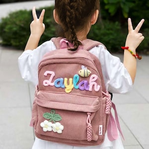 May include: A pink corduroy backpack with the name "Zaylah" in colourful letters. The backpack features a bee and flower embellishments. It has a front pocket, side pocket, and pink shoulder straps.