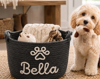May include: A dark gray round woven rope basket featuring a white embroidered paw print and the name Bella in script font. The basket contains a cream-colored knit blanket and a small toy, while a light-colored puppy holds a plush toy nearby.