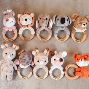 Op de afbeelding: Een verzameling handgemaakte gehaakte babyrammelaars in verschillende dierenvormen, waaronder een hert, leeuw, olifant, koala, beer, vos en koe. Elke rammelaar heeft een houten ring en is gepersonaliseerd met een naam.