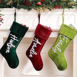 Peut inclure: Trois bas de Noël en velours vert, rouge et vert citron, chacun brodé d'un nom en blanc : "Hudson", "Amelia" et "Emily". Les bas sont suspendus à une cheminée blanche, décorée de verdure festive et de baies rouges.