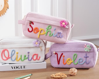 May include: Three rectangular corduroy pencil cases in light pink, white, and lavender. Each case features a personalized name embroidered in colorful yarn: Sophia, Olivia, and Violet. Small yarn flowers are attached to the top right corners.