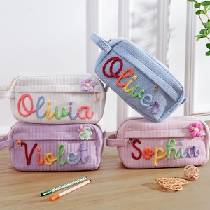 Op de afbeelding: Vier corduroy etuis in pastelkleuren: lichtblauw, roze, lavendel en gebroken wit. Elke etui heeft een naam geborduurd in regenboogkleuren: Olivia, Violet, Sophia en Oliver. Bloemenversieringen en ritssluitingen zijn zichtbaar.