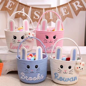 May include: Four Easter baskets in white, pink, and blue, each with bunny ears and a face design. The baskets have handles and embroidered names: Evelyn, Stella, Brooks, and Rowan. A banner saying "Easter" is in the background.