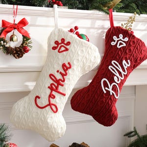 Peut inclure: Deux bas de Noël en forme d'os, un crème et un rouge, suspendus à une cheminée. Le bas crème porte le nom "Sophia" brodé en rouge, et le bas rouge porte le nom "Bella" en blanc. Les deux ont des empreintes de pattes.
