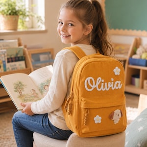 May include: A mustard yellow corduroy backpack featuring the name Olivia in white script lettering, adorned with two white flower patches and a small brown and white puppy dog patch, with braided zipper pulls on the main and front compartments.