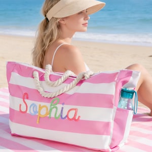 Puede incluir: Un bolso de playa a rayas rosas y blancas con el nombre "Sophia" bordado en hilo colorido. El bolso tiene asas de cuerda y un bolsillo lateral que contiene una botella de agua. El bolso está sobre una toalla de playa a rayas rosas y blancas.