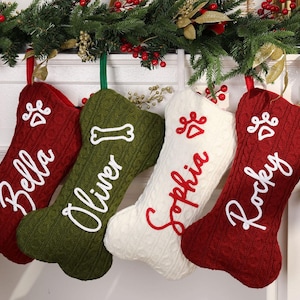 May include: Four personalised Christmas stockings in red, green, and white, each with a pet's name in cursive. The stockings are decorated with paw prints and a bone, hanging from a garland of greenery and berries.