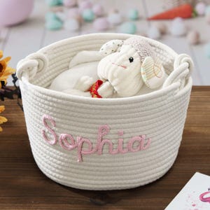 K&ouml;nnte beinhalten: Ein runder, cremewei&szlig;er Korb aus geflochtenem Seil mit dem Namen Sophia, der in rosa Garn auf der Vorderseite eingestickt ist. Der Korb hat zwei integrierte Seilgriffe an den Seiten und enth&auml;lt ein wei&szlig;es Pl&uuml;schschaf mit einem roten Band.