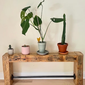May include: A rustic wooden bench with a black metal bar underneath. Three potted plants are displayed on the bench, including a cactus, a succulent, and a monstera plant.