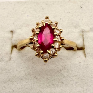 May include: A gold ring featuring a central marquise-cut deep pink gemstone surrounded by a halo of small, clear, round-cut stones. The setting is crafted from yellow gold, with the central stone held in place by multiple prongs.