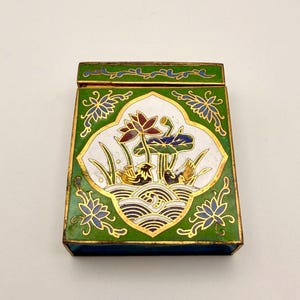 May include: A rectangular metal matchbox cover featuring green enamel with gold-toned metal outlines. The center displays a white oval panel with a colorful depiction of two birds, lotus flowers, and stylized water waves. The lid has a vine pattern.