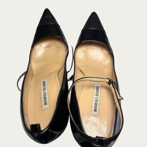 May include: Pair of black patent leather pumps with pointed toes. The shoes have a Mary Jane strap and the brand name "MANOLO BLAHNIK" is visible on the insole. The shoes are on a white background.