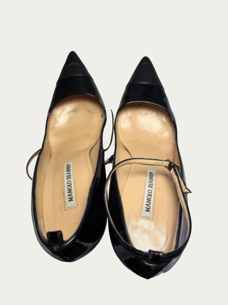 May include: Pair of black patent leather pumps with pointed toes. The shoes have a Mary Jane strap and the brand name "MANOLO BLAHNIK" is visible on the insole. The shoes are on a white background.