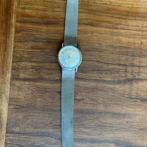 May include: A silver-toned wristwatch with a mesh band lies on a wooden surface. The watch face is round with a light-colored dial and silver hands. The band is a fine, woven mesh, and the watch appears to be vintage.