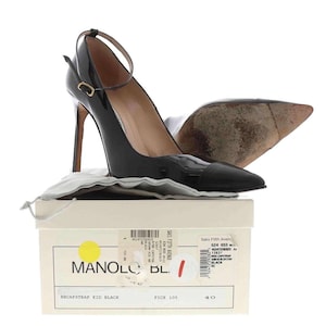 May include: Black patent leather high-heeled pumps with ankle straps. The shoes have pointed toes and are displayed on a box with the brand name "MANOLO" and the text "BBCAPSTRAP KID BLACK". The box is white with a yellow circle.