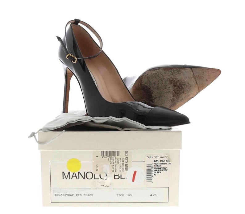 May include: Black patent leather high-heeled pumps with ankle straps. The shoes have pointed toes and are displayed on a box with the brand name "MANOLO" and the text "BBCAPSTRAP KID BLACK". The box is white with a yellow circle.