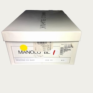 May include: A white shoe box with the brand name "MANOLO BLAHNIK" printed on the top. The box has a yellow sticker and a label with the text "BBCAPSTRAP KID BLACK" and size "40".