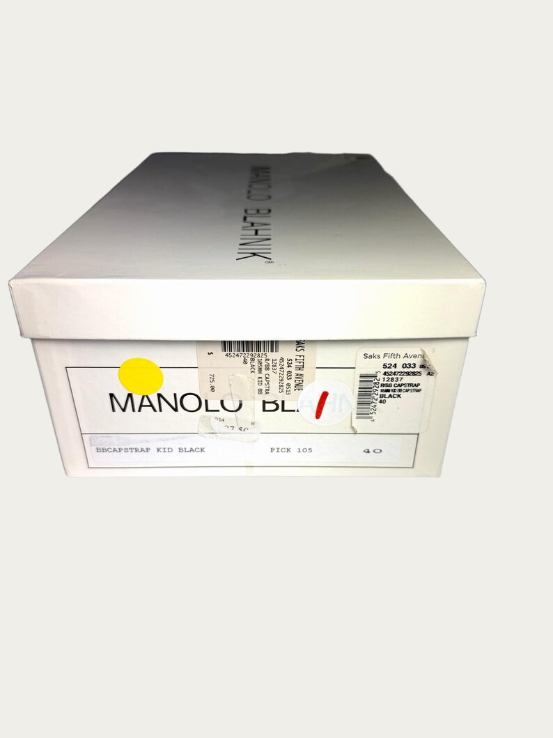 May include: A white shoe box with the brand name "MANOLO BLAHNIK" printed on the top. The box has a yellow sticker and a label with the text "BBCAPSTRAP KID BLACK" and size "40".