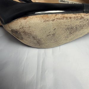 May include: Close-up of a black high-heeled shoe with a worn sole. The shoe has a glossy finish and a strap. The sole shows signs of wear and tear, with visible scuff marks and discoloration. The shoe is set against a white background.