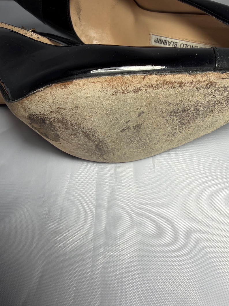May include: Close-up of a black high-heeled shoe with a worn sole. The shoe has a glossy finish and a strap. The sole shows signs of wear and tear, with visible scuff marks and discoloration. The shoe is set against a white background.
